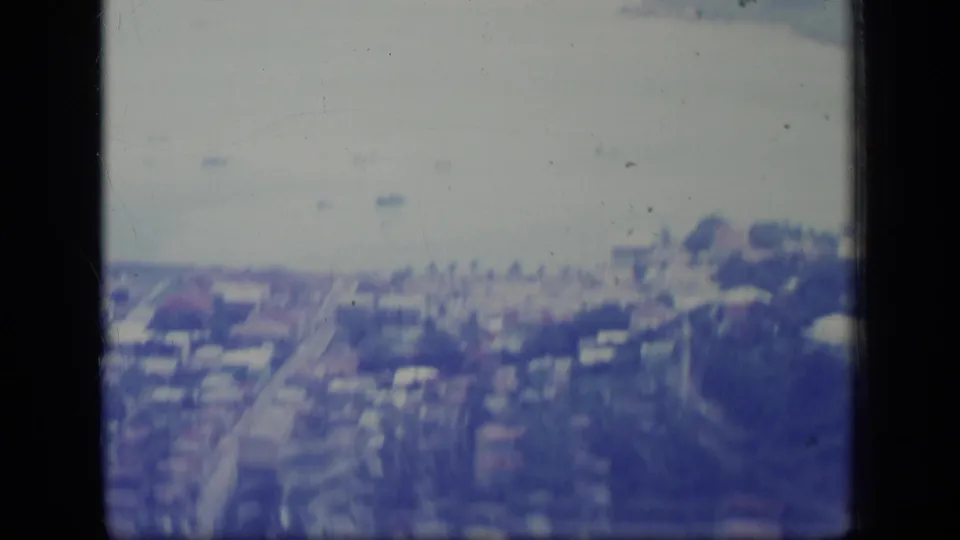 Soft 8mm aerial view in Puerto Rico, with coastal buildings, a road grid, pale water, and small boats in the distance.