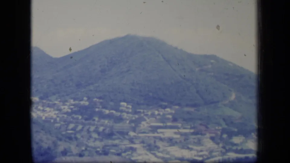1970 Puerto Rico aerial frame showing a prominent green mountain above low buildings and open foreground land.