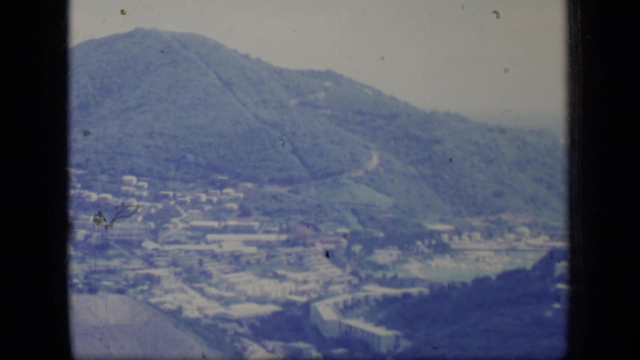 Distant mountain town below green hills with roads and pale buildings under haze.