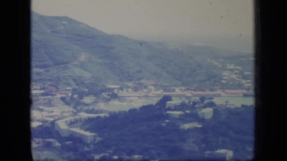 Soft 8mm frame of Puerto Rico foothills in 1970, showing hillside vegetation, valley roads, open ground, and distant haze.