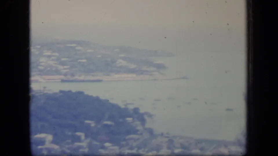 Soft 1970 Puerto Rico 8mm frame with shoreline development, pale bay water, wooded foreground, and distant coastal land.