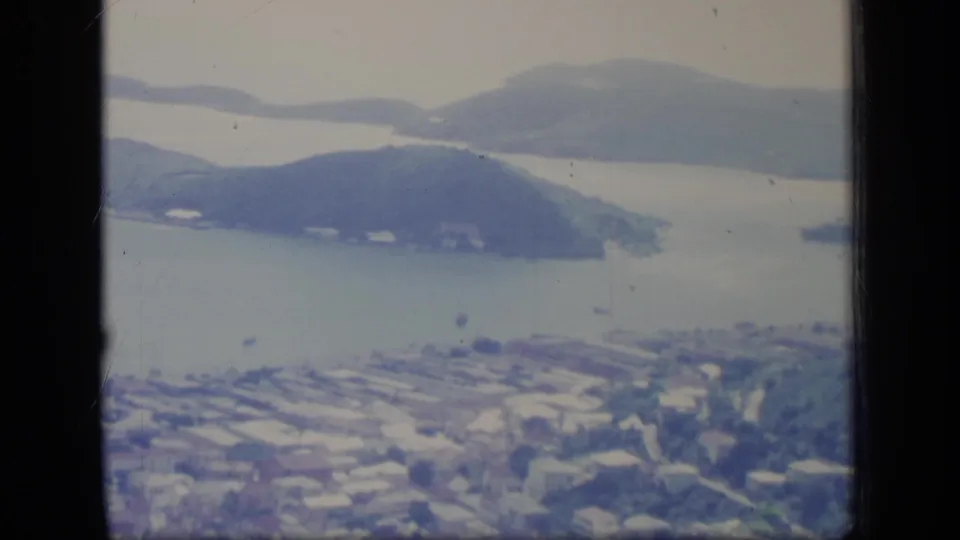 1970 Puerto Rico harbor view from above, with foreground town rooftops, small boats, rounded islands, and hazy hills across the water.