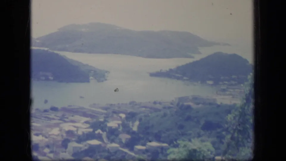 1970 Puerto Rico home-movie frame showing a sheltered bay, rounded green hills, shoreline buildings, and soft foreground vegetation.