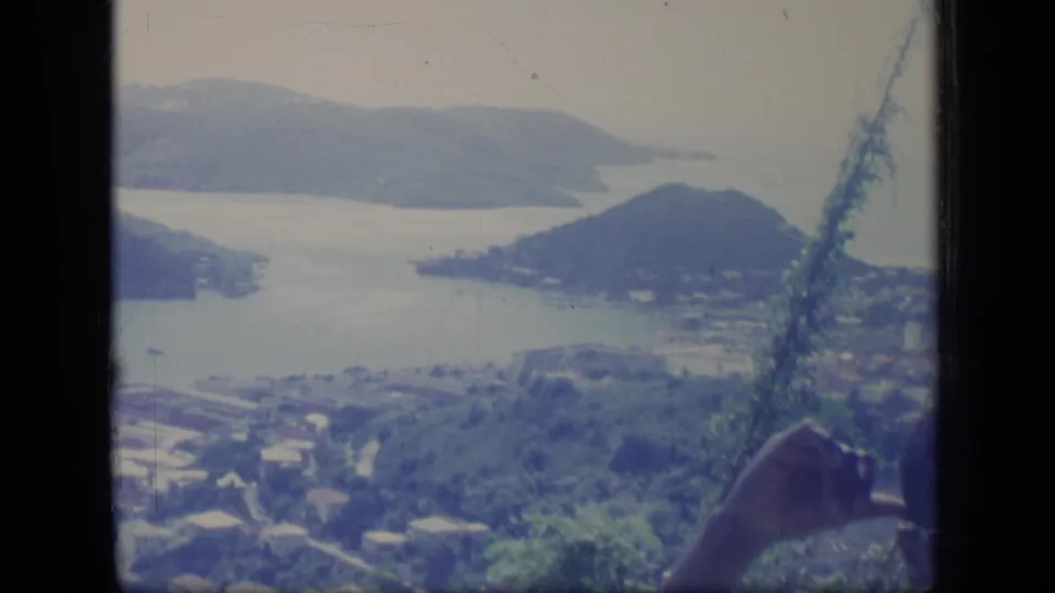 1970 Puerto Rico 8mm frame of a coastal bay and hills, with foreground vegetation and a person's hand entering at lower right.