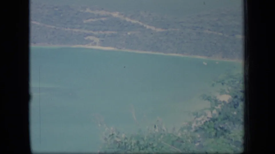 1970 Puerto Rico aerial home-movie frame showing turquoise coastal water, a narrow beach, and wooded land through a window or film gate edge.