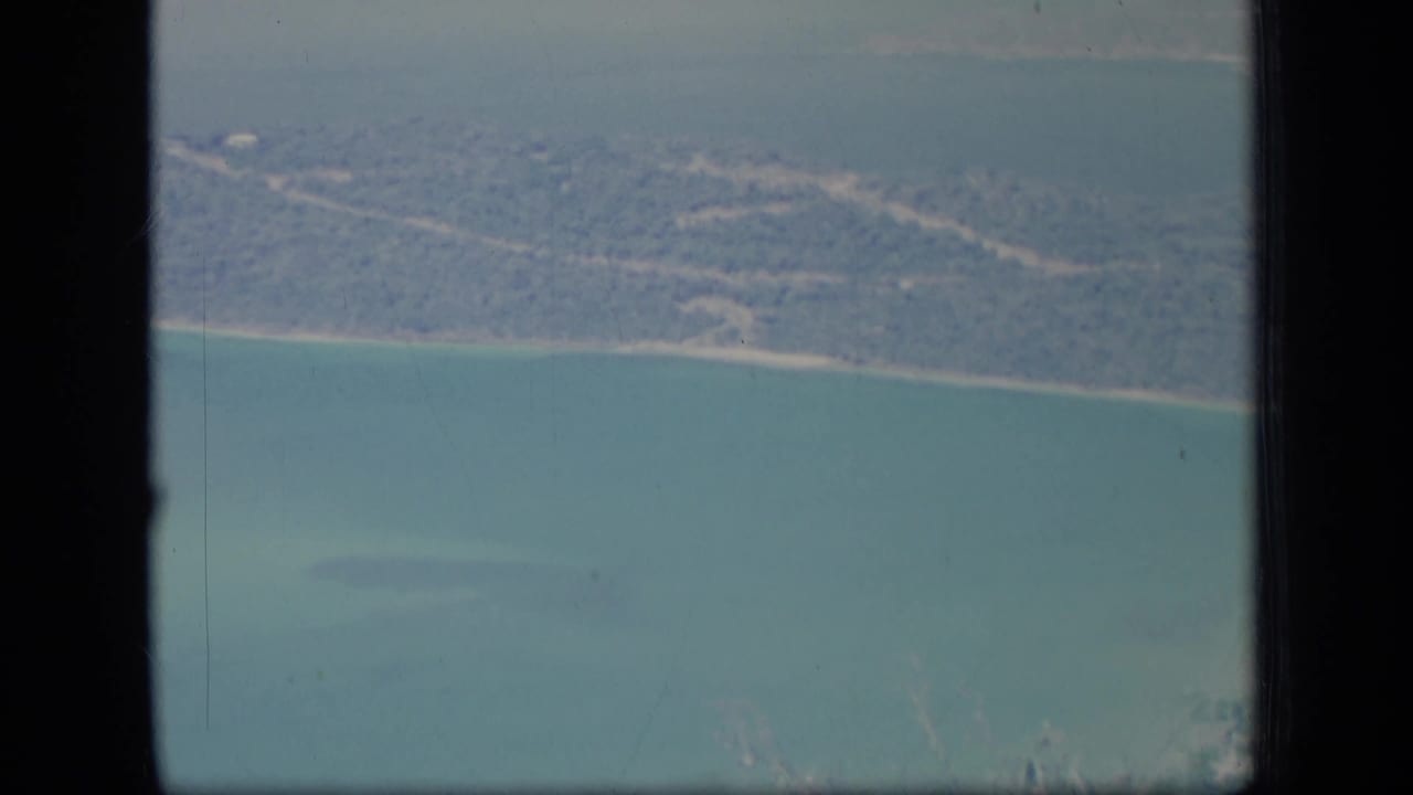Aerial 8mm view of Puerto Rico coastline in 1970, with pale shore, wooded land, turquoise water, and dark window framing.