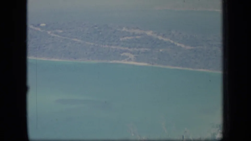 Aerial 8mm view of Puerto Rico coastline in 1970, with pale shore, wooded land, turquoise water, and dark window framing.