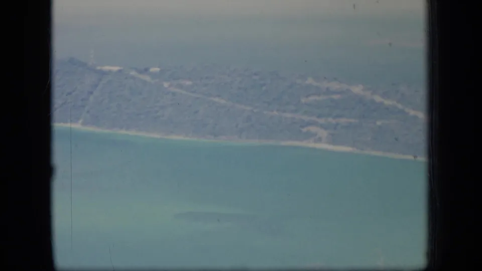 1970 Puerto Rico home-movie frame showing an aerial pass over wooded coastal hills, a beach line, and turquoise water.