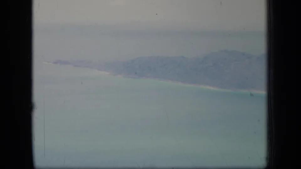 1970 Puerto Rico home-movie frame showing a distant aerial view of green coastal land, pale beach, and turquoise water with visible film grain and border.