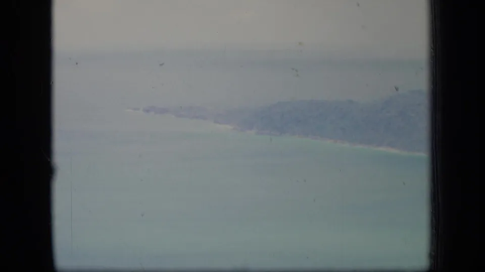 1970 Puerto Rico 8mm frame showing a hazy aircraft-window view of ocean, pale sky, and a distant forested coastline.