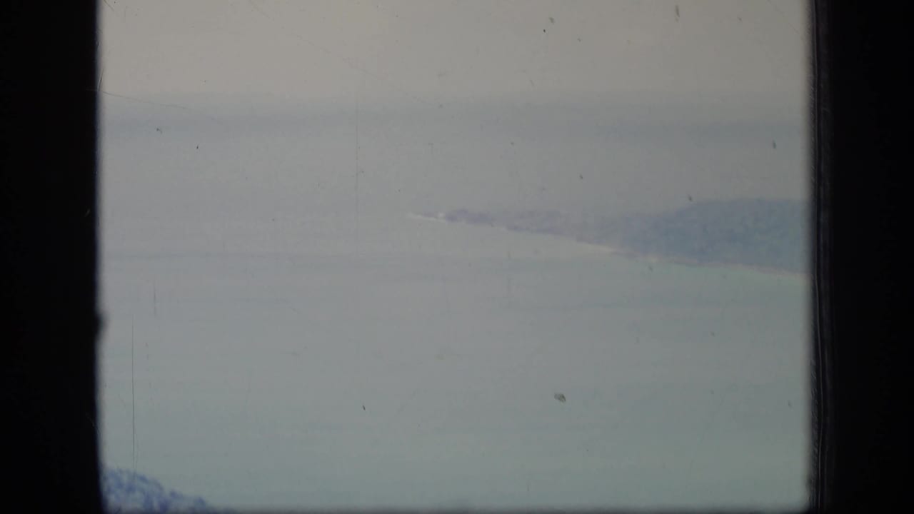 Soft aerial view of ocean and distant wooded shore partly framed by vertical aircraft-window borders.