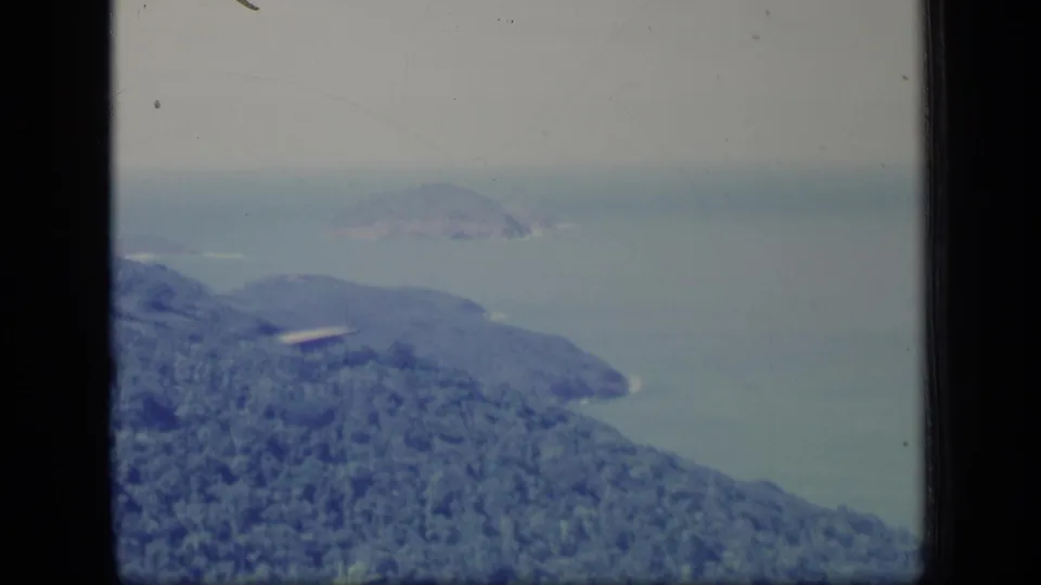1970 Puerto Rico aerial home-movie frame showing a wooded coastal headland, blue-green water, haze, and film wear.