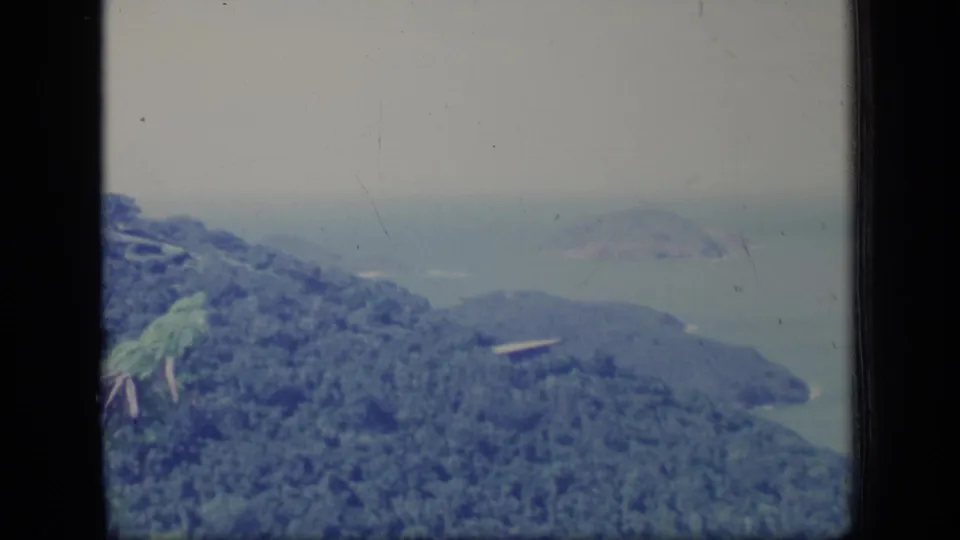 1970 Puerto Rico 8mm frame of a wooded coastal slope above hazy sea, with a small pale structure visible on the ridge.