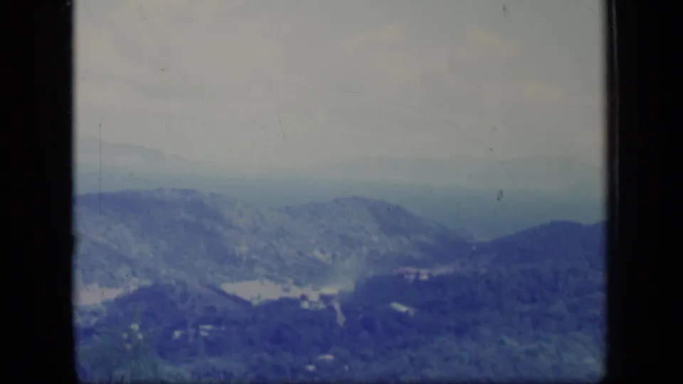 A 1970 Puerto Rico 8mm frame showing forested mountains, a light valley clearing, and scattered small buildings beneath a hazy partly cloudy sky.