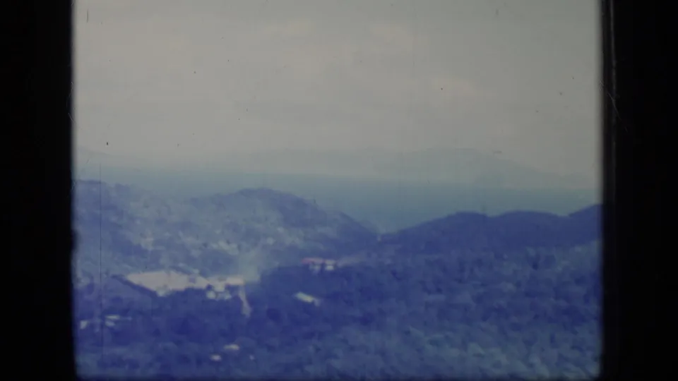 A faded 8mm view from 1970 Puerto Rico of blue-toned wooded hills, a valley clearing, and scattered buildings seen from a high or airborne vantage point.
