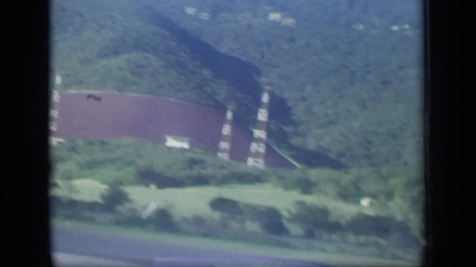 Aerial Puerto Rico frame showing a red-roofed structure, tall communication towers, roads, and green mountain terrain.