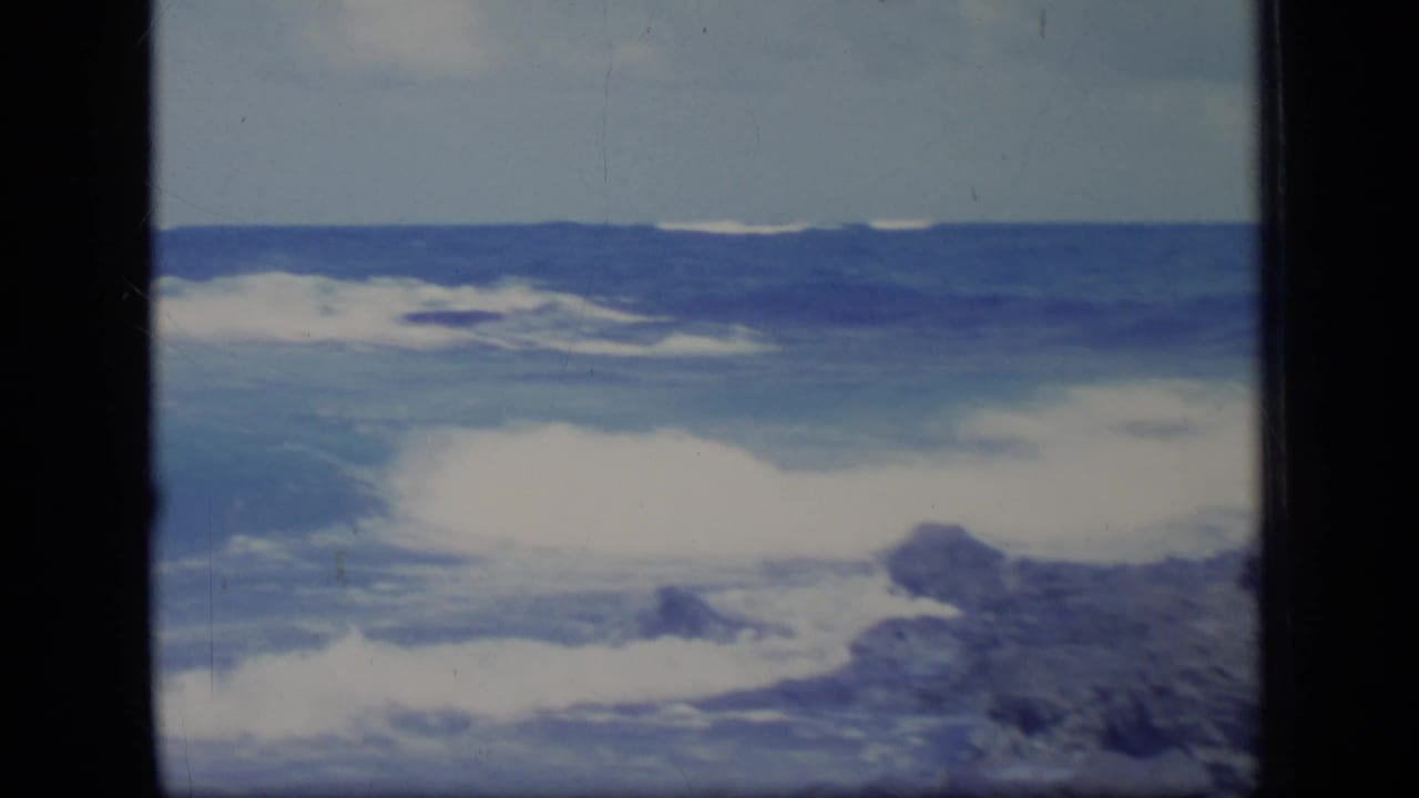 1970 Puerto Rico coastal frame with blue water, white surf, and dark shoreline rocks under a bright sky.