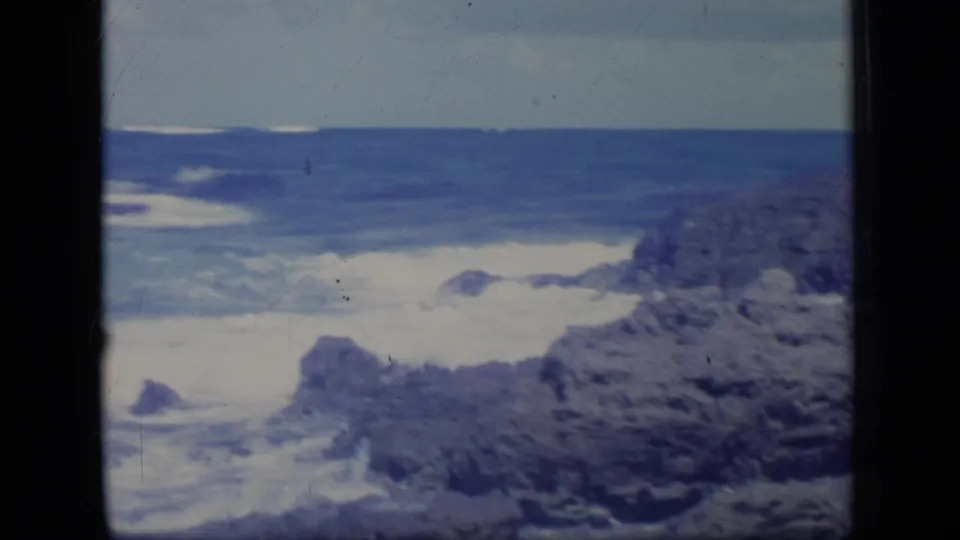1970 Puerto Rico 8mm frame showing foamy surf washing around a rocky shoreline with open blue water beyond.