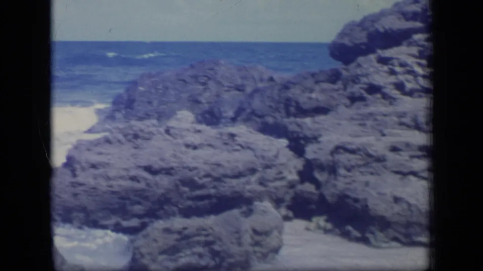1970 Puerto Rico coastal frame with large dark rocks in the foreground and breaking waves across the blue ocean behind them.