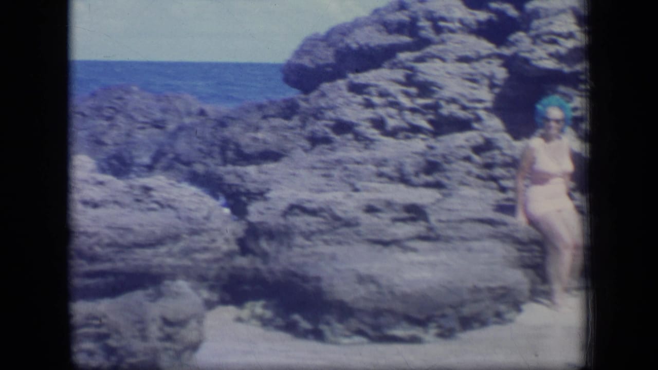 Woman in a light swimsuit stands near rugged coastal rocks with ocean behind her.