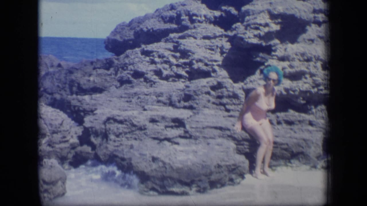 Woman stands on pale sand beside dark rocks as waves approach the shore.