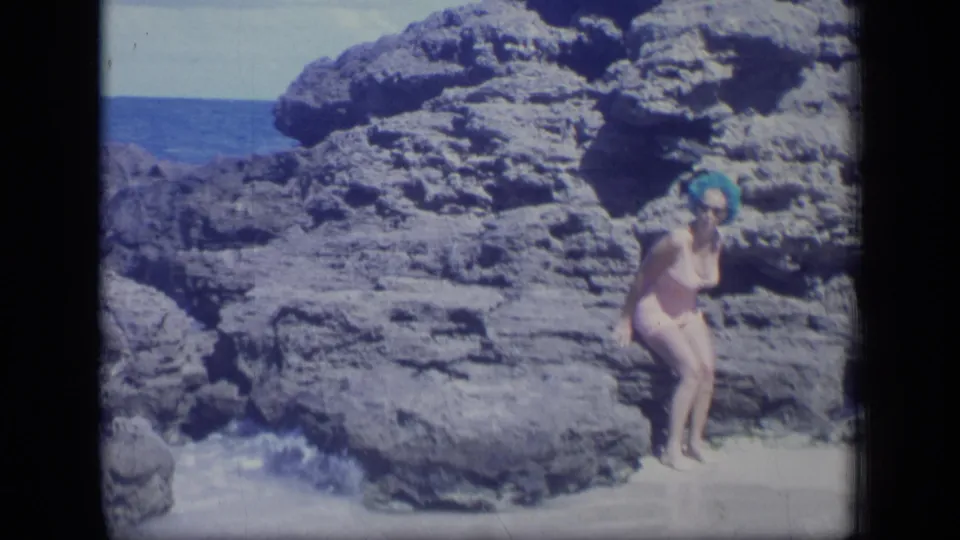 1970 Puerto Rico frame of a woman in a one-piece swimsuit standing at the edge of coastal rocks near incoming surf.