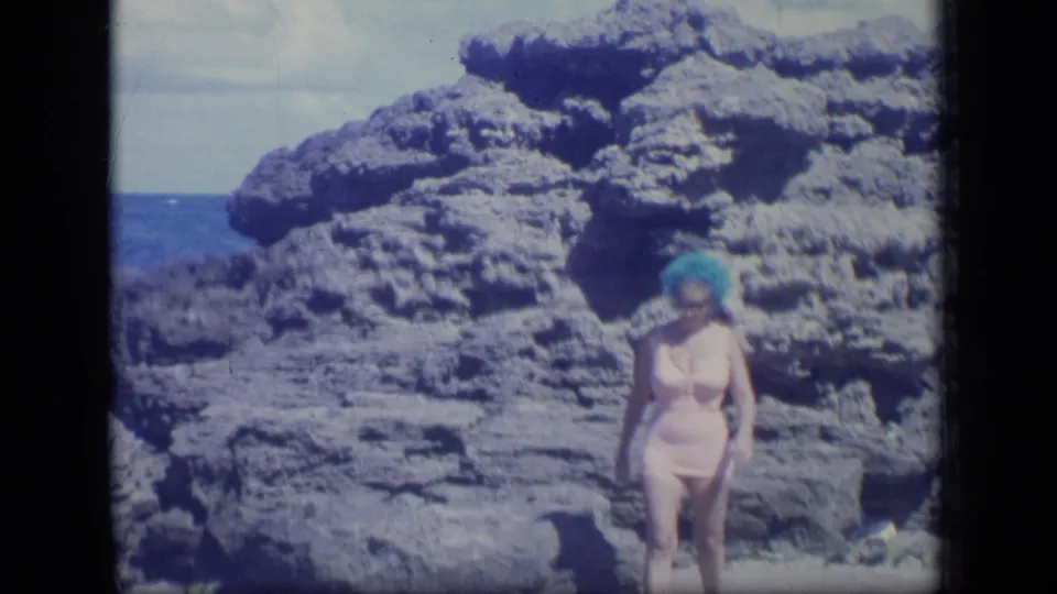 1970 Puerto Rico frame with the woman approaching the camera, backed by rugged shoreline rocks and a strip of blue ocean.