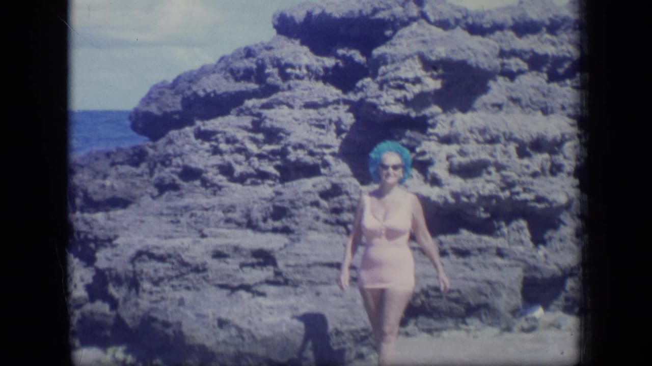 Woman in a pink swimsuit walks through shallow water with large rocks behind her.