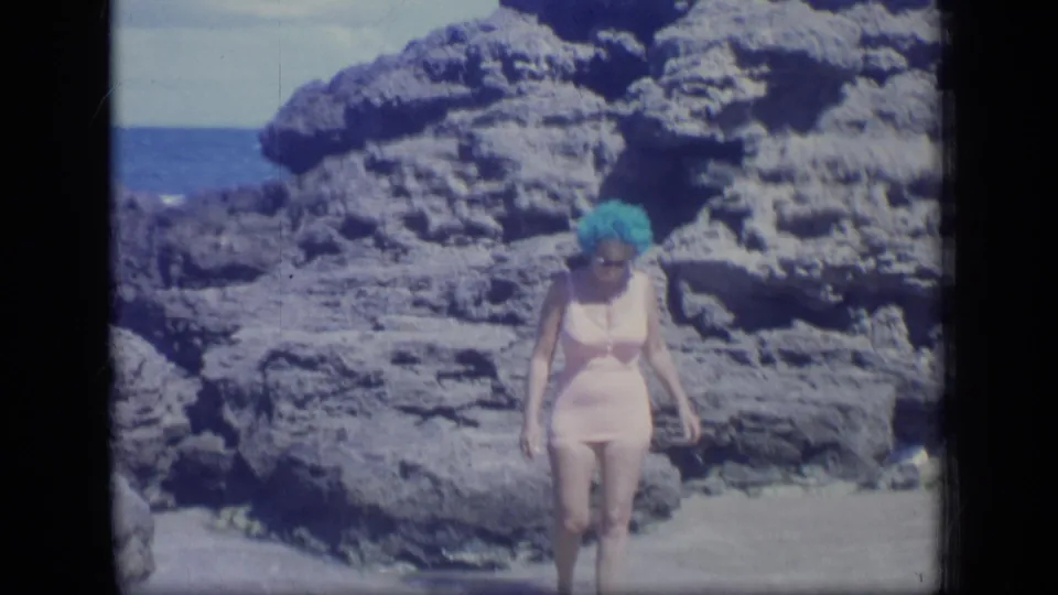 The woman continues across shallow water on a rocky Puerto Rico beach, with the ocean visible beyond the stone formations.