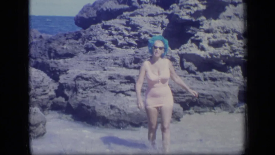 A woman in sunglasses and a vintage pink swimsuit walks toward the camera through shallow beach water in Puerto Rico.