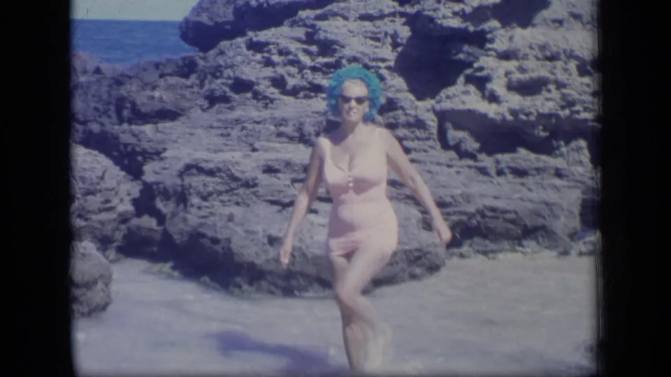 At the end of the clip, the woman steps across shallow water in front of rugged coastal rocks and blue ocean.