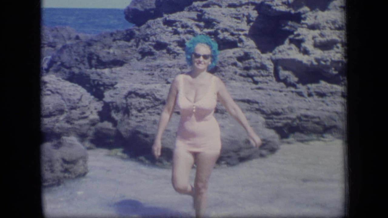 Woman in a pink swimsuit and blue headwear walks on sand near dark rocks.
