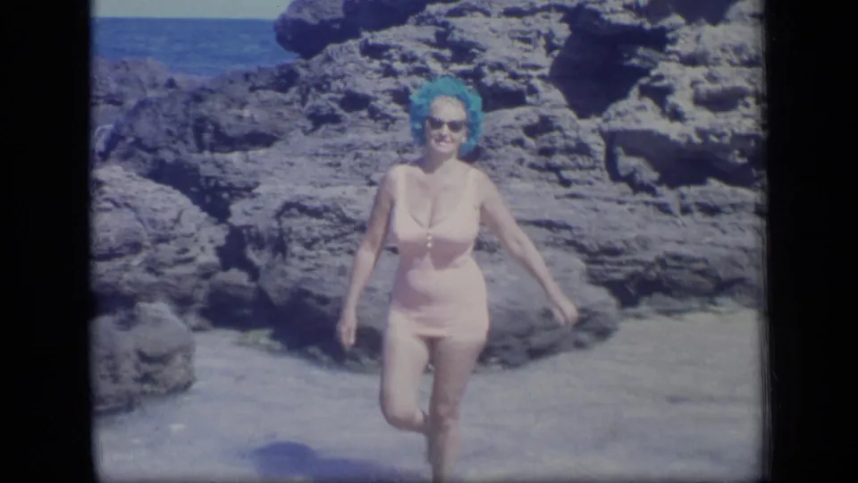 Adult woman in a pink one-piece swimsuit, blue wig or swim cap, and sunglasses walks along a rocky Puerto Rico beach in 1970 home-movie footage.