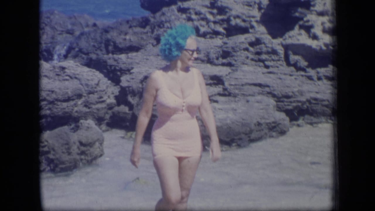 Adult woman in pink swimwear and bright blue headwear walks across sand beside rugged rocks on a Puerto Rico beach.
