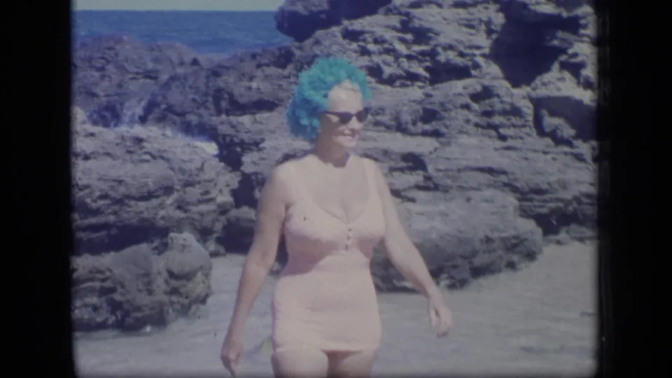 Smiling adult woman in a pink one-piece swimsuit, sunglasses, and bright blue headwear walks near volcanic-looking beach rocks in Puerto Rico.