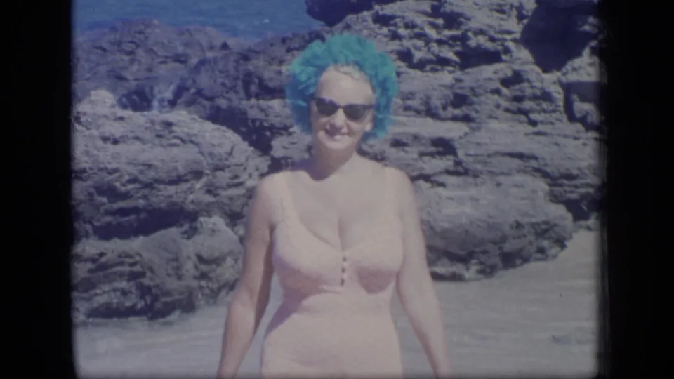 Closer 1970 home-movie view of an adult woman in a pink swimsuit, sunglasses, and blue wig or swim cap standing near rocks on a Puerto Rico beach.