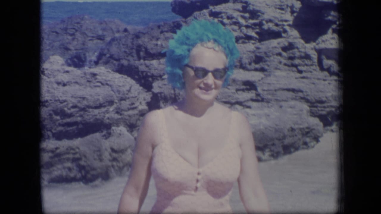 In Puerto Rico home-movie footage from 1970, a woman in a turquoise swim cap and sunglasses smiles near coastal rocks and water.