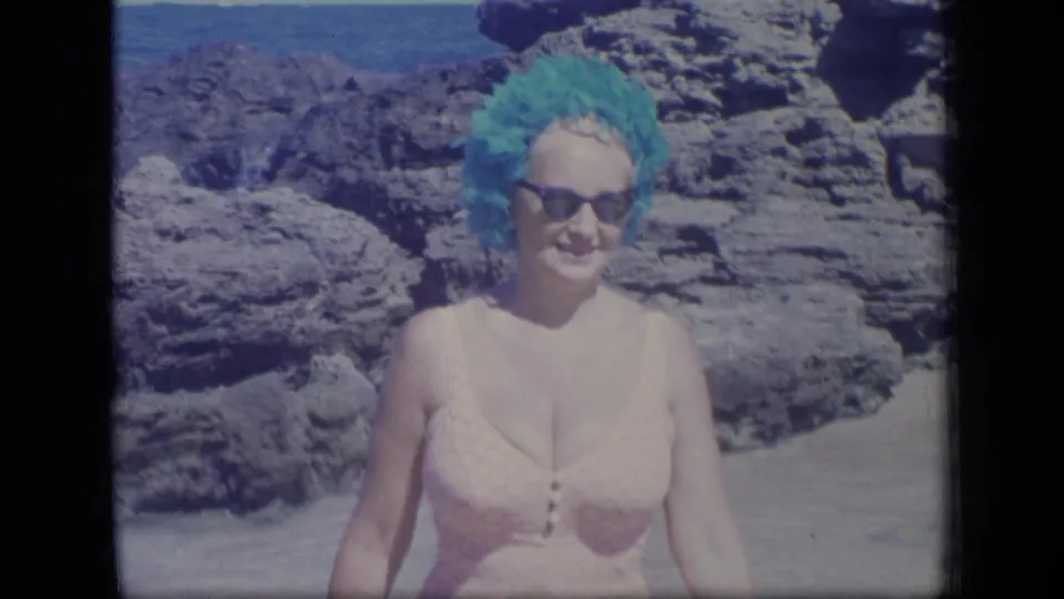 In Puerto Rico home-movie footage from 1970, a woman in a turquoise swim cap and sunglasses smiles near coastal rocks and water.