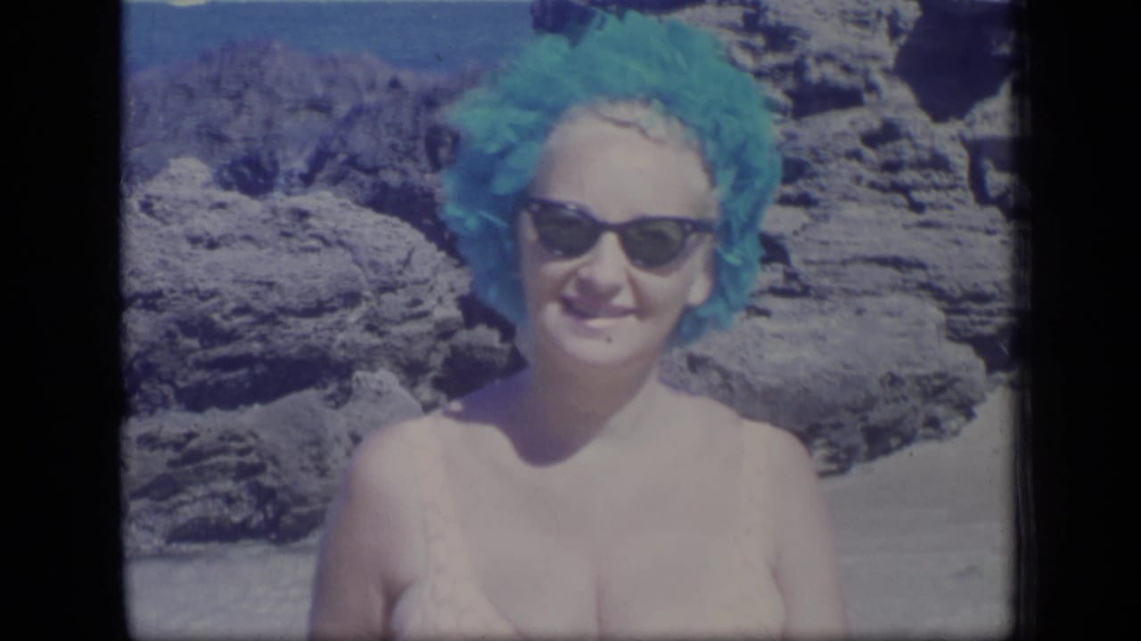 Closer 1970 Puerto Rico beach view of a smiling woman wearing sunglasses and a turquoise swim cap, with rocks and ocean behind her.