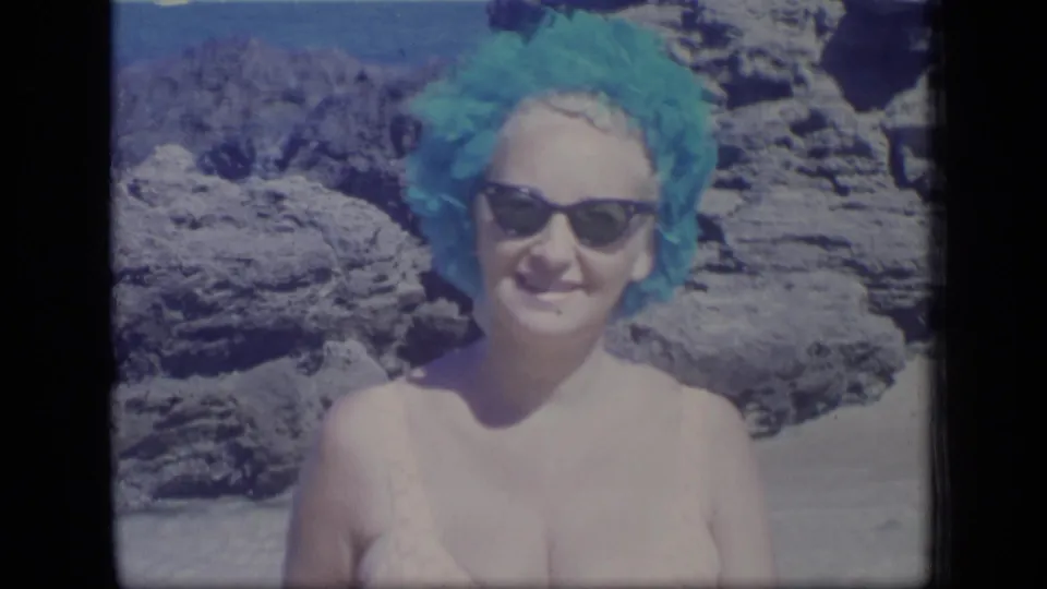 Closer 1970 Puerto Rico beach view of a smiling woman wearing sunglasses and a turquoise swim cap, with rocks and ocean behind her.