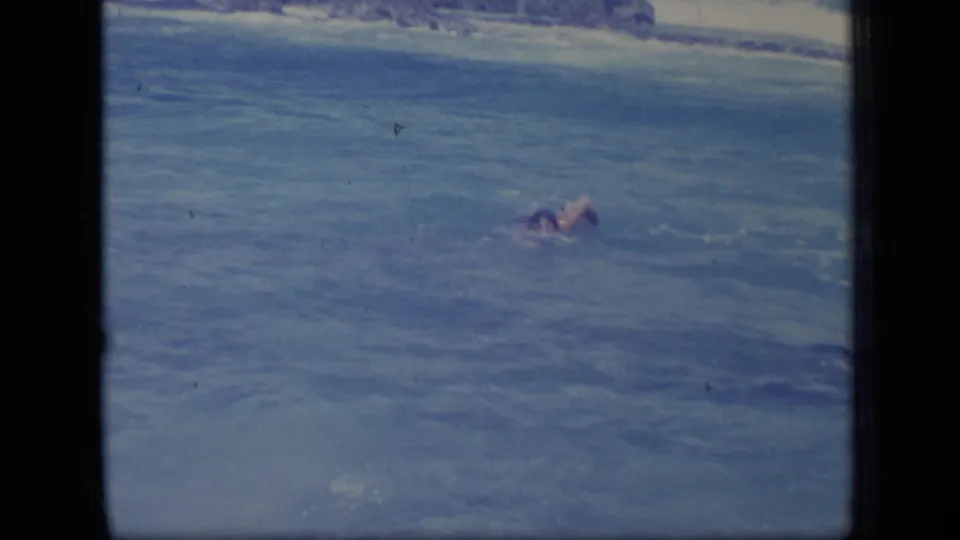 A distant swimmer moves through blue coastal water in the same 1970 Puerto Rico home-movie beach sequence.