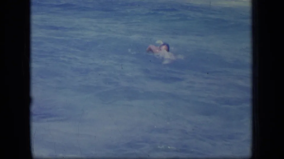 In 1970 Puerto Rico footage, a man swims in blue ocean water, seen at a distance within dark 8mm gate borders.