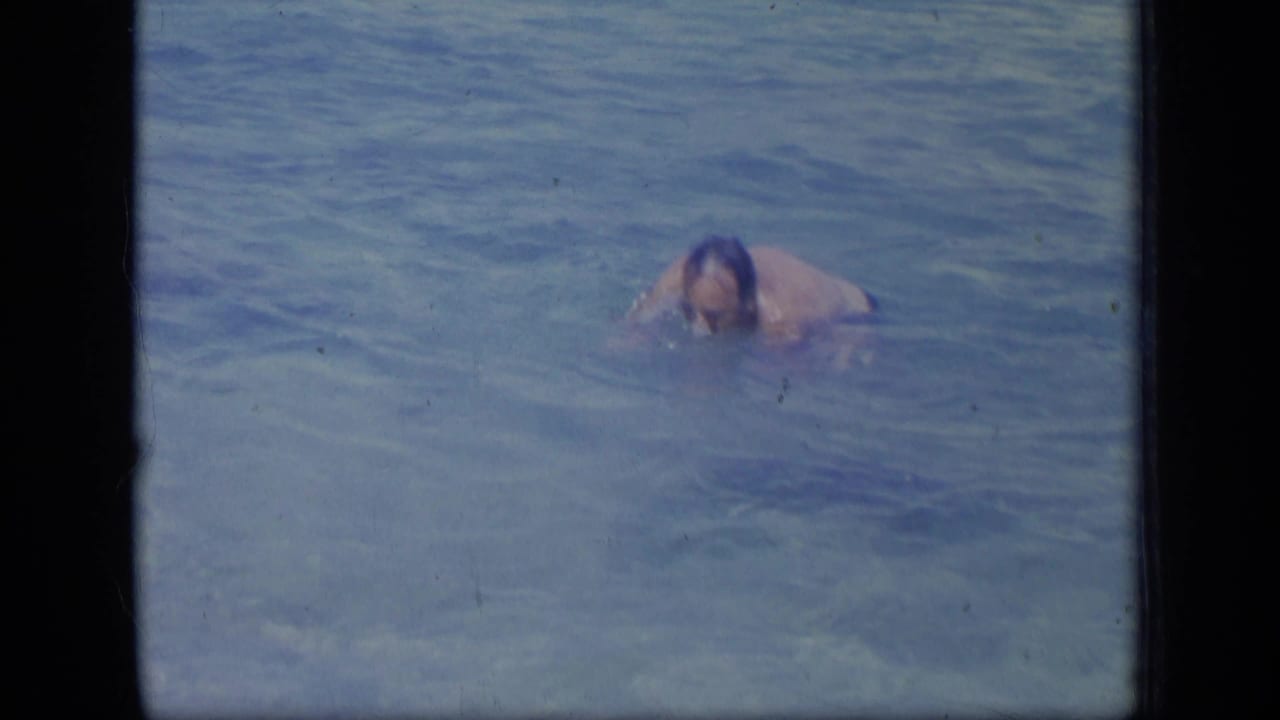 In 1970 Puerto Rico home-movie footage, an adult man is mostly submerged in shallow blue ocean water.