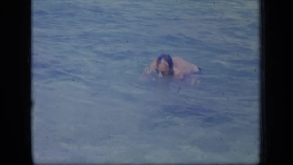 In 1970 Puerto Rico home-movie footage, an adult man is mostly submerged in shallow blue ocean water.