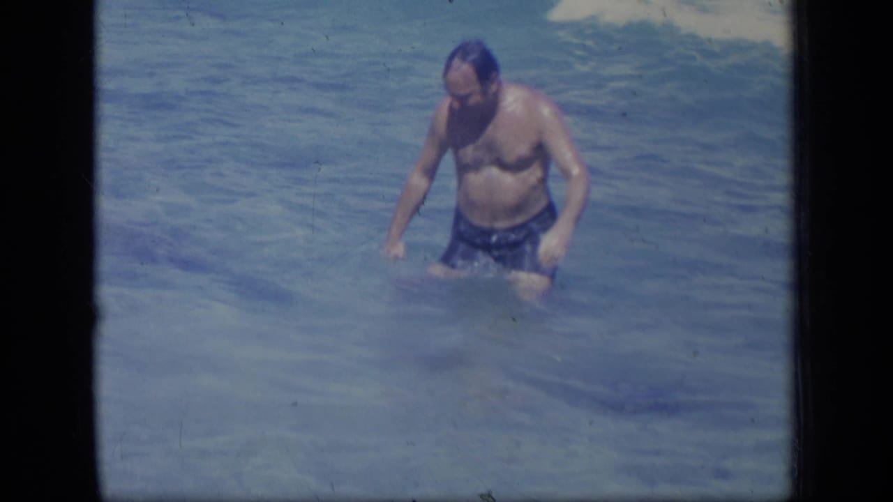 Adult man in dark swim shorts standing in shallow blue water.