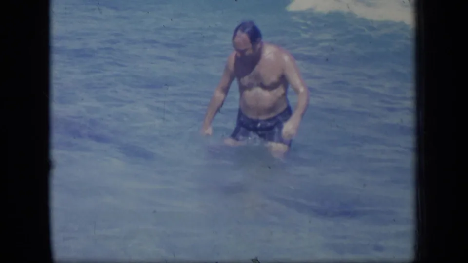 The man stands waist-deep in the ocean, wearing dark swim shorts in the supplied 1970 Puerto Rico setting.