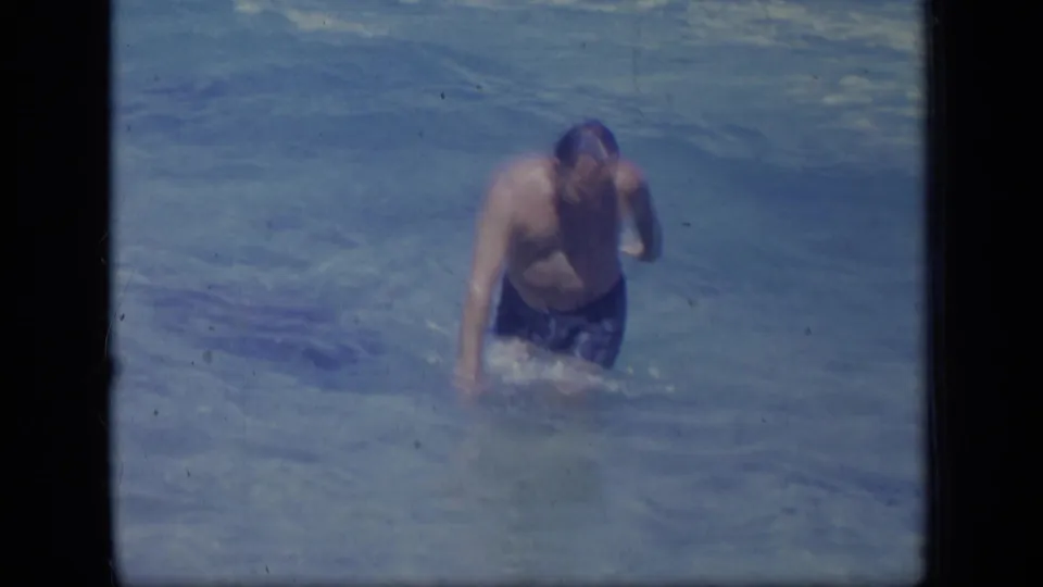The man moves through the shallow ocean water, splashing lightly while one hand is raised near his head.