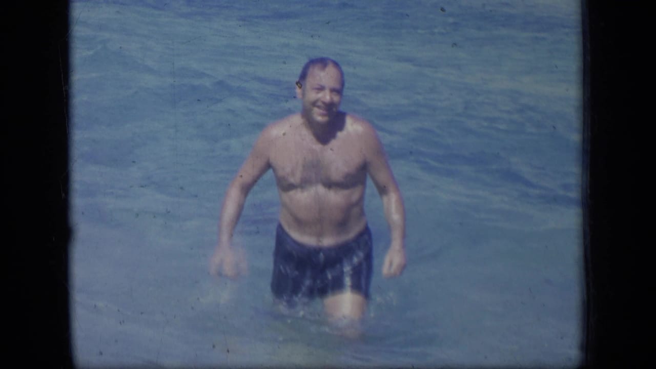 Adult man in blue swim trunks smiles while standing in clear shallow water.