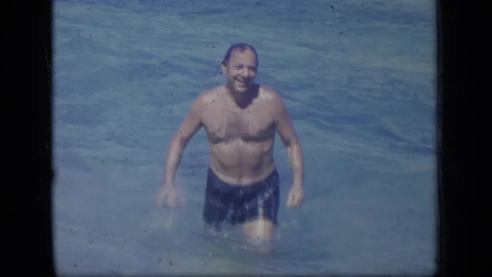 In 1970s Puerto Rico home-movie footage, an adult man in blue swim trunks smiles toward the camera while wading in clear shallows.