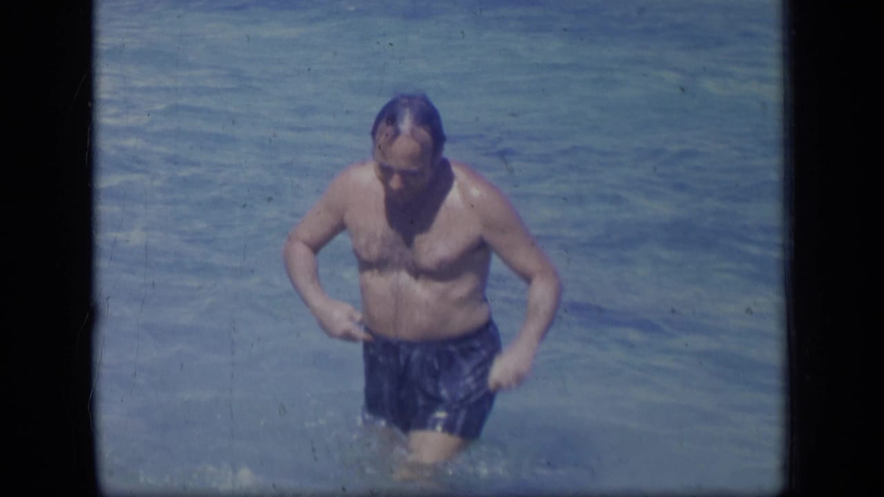 Adult man looks down while adjusting blue swim trunks in shallow water.